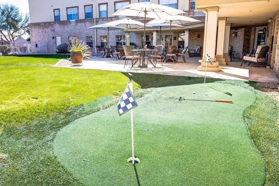 Outdoor putting green with two golf holes and flags in front of a covered patio area with tables, chairs, and umbrellas at an assisted living facility.