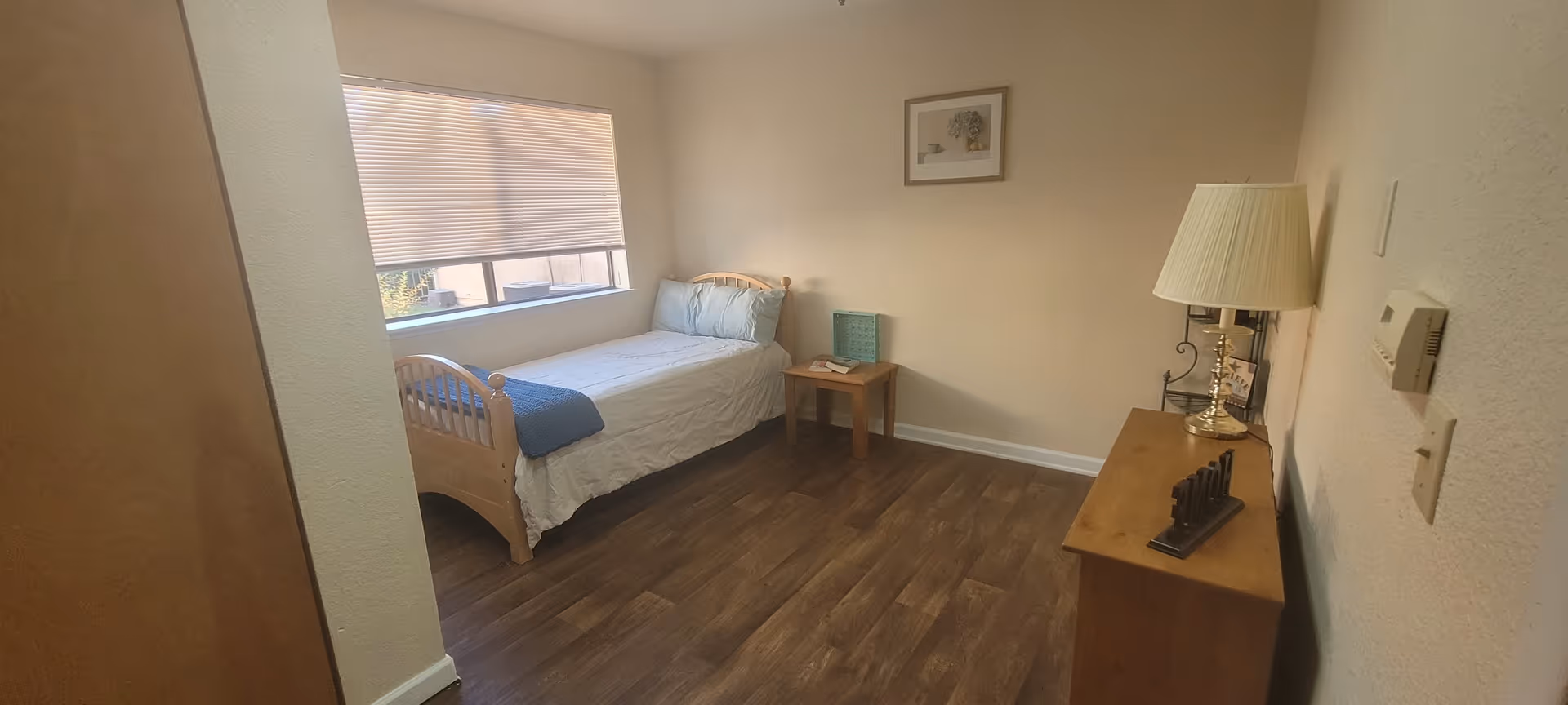 A small bedroom with a single wooden bed against the wall under a window with closed blinds. The bed is made with white sheets, a blue blanket, and two pillows. Next to the bed is a small wooden side table with a book and a decorative item. On the opposite wall, there is a wooden dresser with a brass lamp and a small decorative piece. The room has wooden flooring and beige walls with a framed picture hanging above the bed.