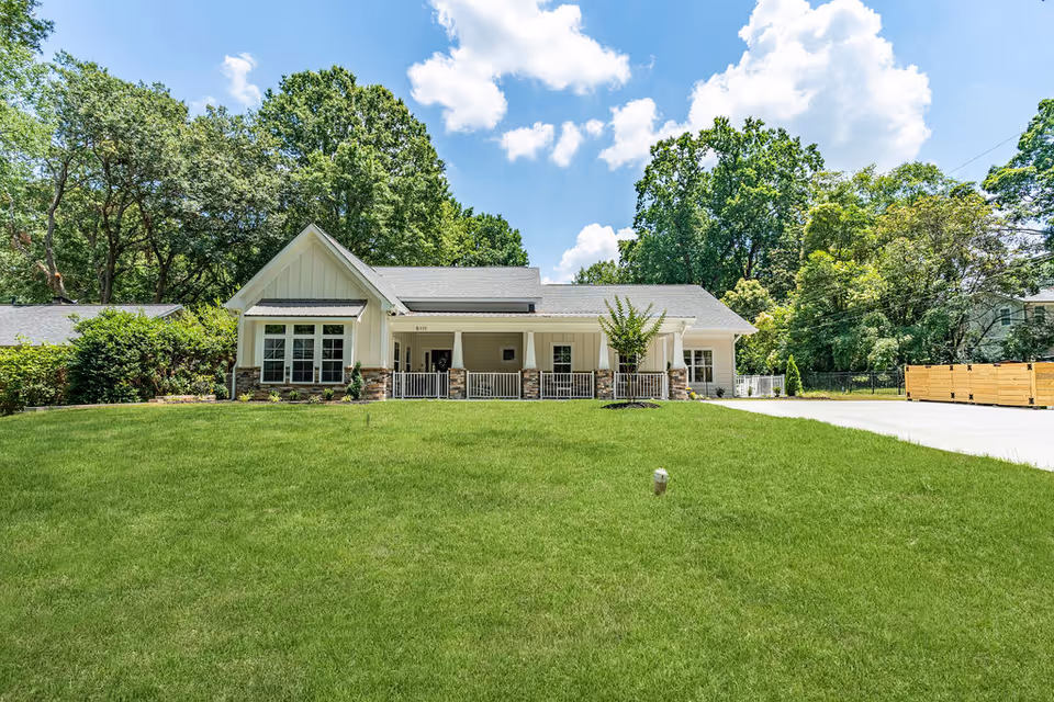 Front view of a single-story residential-style building with a covered porch, a large green lawn, driveway, and trees in the background.