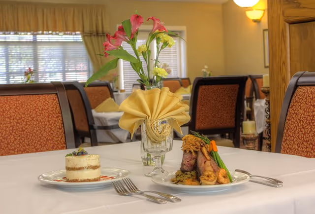 A dining table set with a white tablecloth, a glass with a folded yellow napkin, a vase with pink and yellow flowers, a plated gourmet meal with meat and vegetables, and a dessert on a separate plate. The background shows upholstered dining chairs and windows with blinds in a warmly lit dining room.