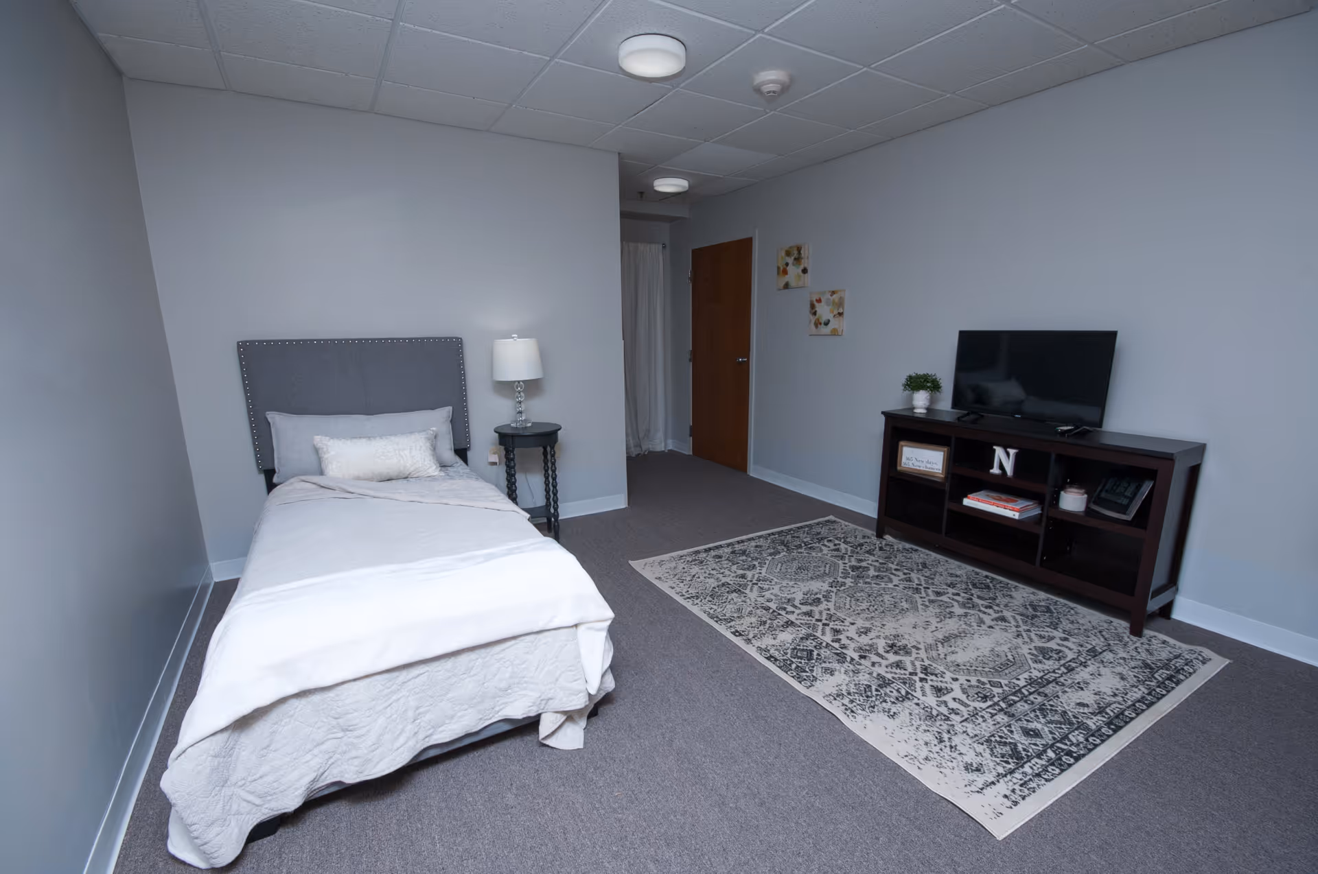A simple senior living bedroom with a single bed featuring a gray upholstered headboard, white bedding, and a small decorative pillow. Next to the bed is a round black side table with a lamp. Across from the bed is a dark wooden TV stand with a flat-screen TV, some books, a plant, and decorative items. The room has gray walls, a patterned area rug on the carpeted floor, and two small framed pictures on the wall near a wooden door.