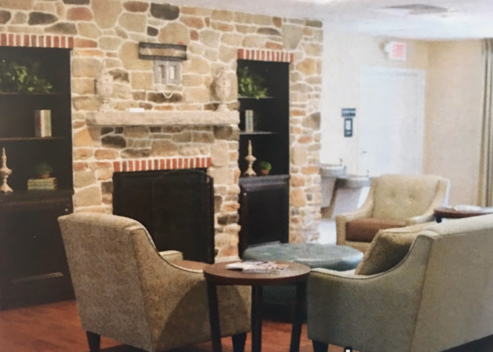 Cozy sitting area with upholstered armchairs and a small round table in front of a stone fireplace with built-in shelves on either side, decorated with books and plants. A water fountain and an exit door are visible in the background.