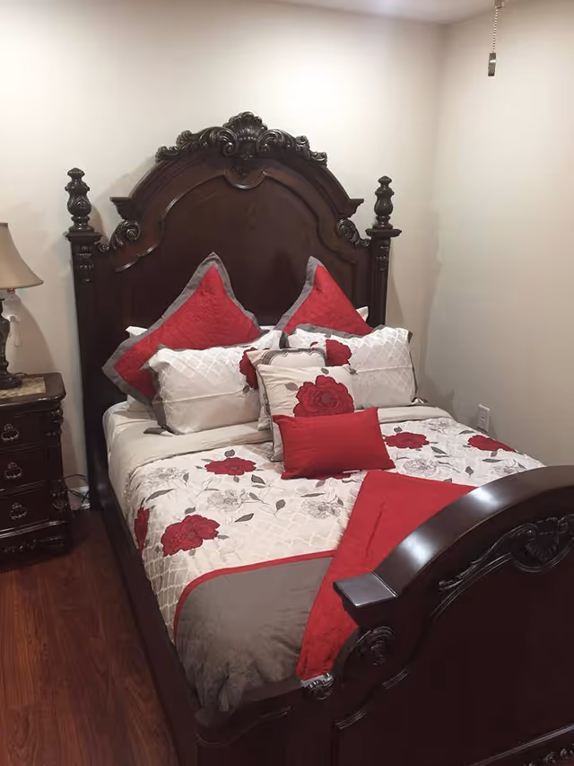 A bedroom with a dark wooden ornate bed frame and headboard. The bed is made with a floral patterned comforter featuring red and gray flowers, and multiple pillows in red, white, and gray colors. There is a wooden nightstand with a lamp on the left side of the bed. The room has wooden flooring and plain light-colored walls.