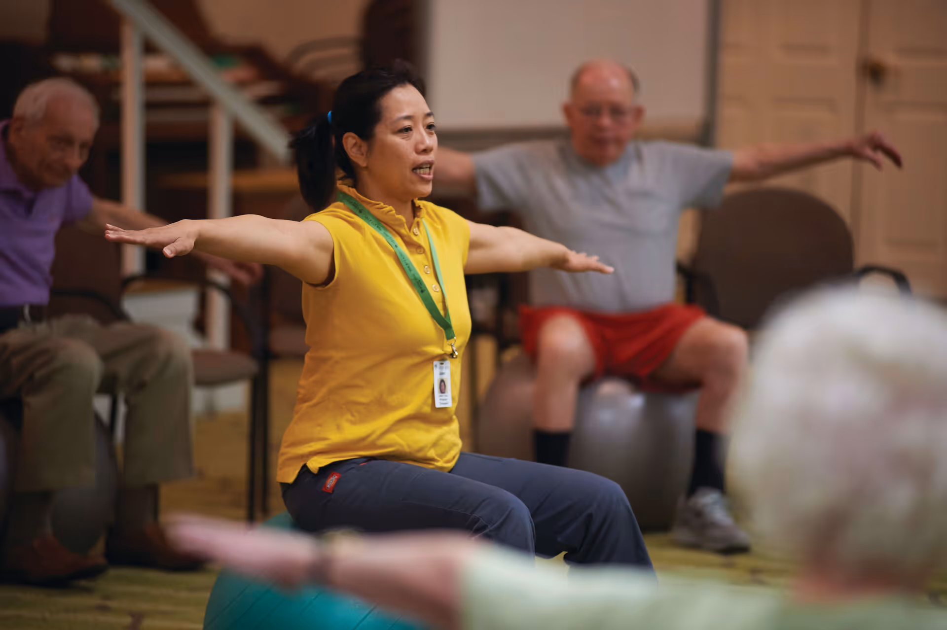 A group of elderly people participating in a seated exercise class led by an instructor wearing a yellow shirt and a green lanyard. They are sitting on exercise balls with their arms extended out to the sides in a spacious indoor room.