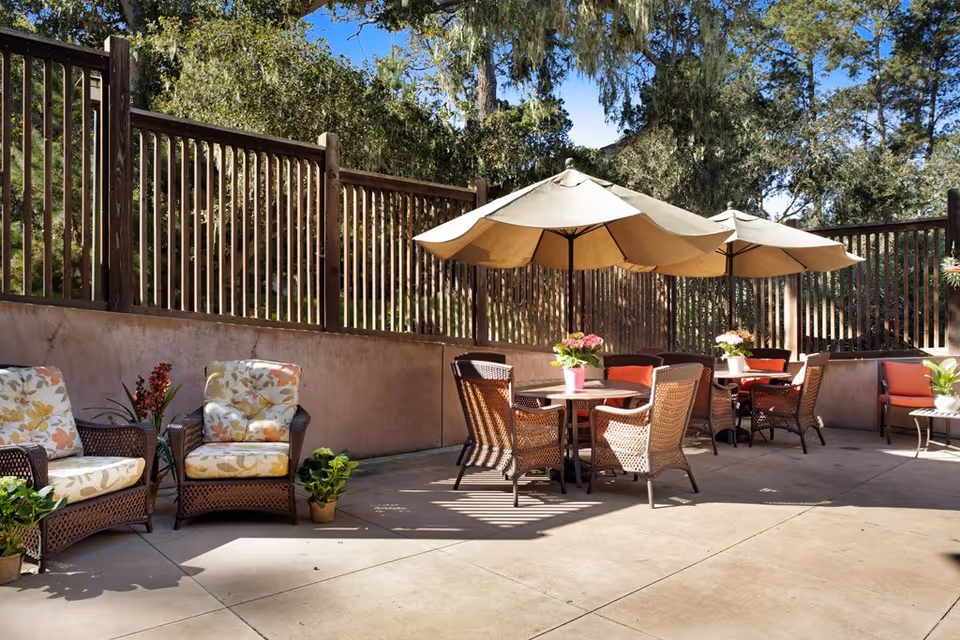 Outdoor patio area with wicker chairs and tables under large beige umbrellas. The chairs have cushions with floral and solid patterns. Potted plants and flowers decorate the space, and a wooden fence with trees is visible in the background under a clear blue sky.