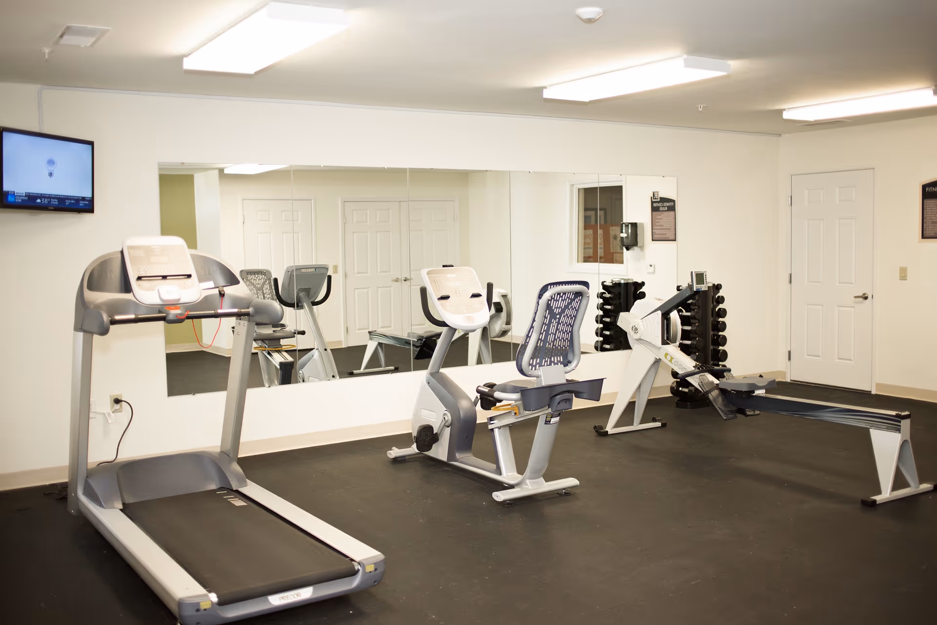 A fitness room with exercise equipment including a treadmill, a recumbent stationary bike, a rowing machine, and a rack of dumbbells. There is a large mirror on the wall reflecting the equipment and a wall-mounted TV in the upper left corner. The room has white walls and a black rubber floor.