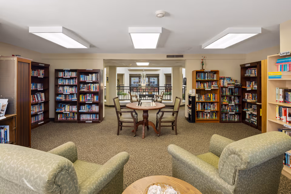 A cozy library room in a senior living community featuring multiple bookshelves filled with books, a round table with four chairs in the center, and two comfortable armchairs in the foreground. The room is well-lit with ceiling lights and has a carpeted floor.