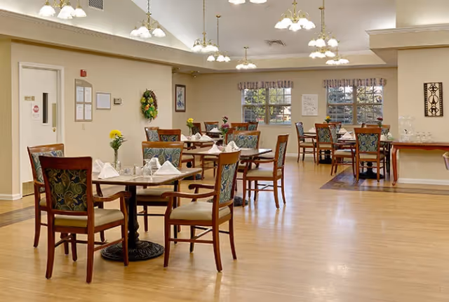 A spacious dining room with several wooden tables and chairs arranged neatly. Each table is set with white napkins, glassware, and small floral centerpieces. The room has large windows with valances, light-colored walls, and multiple ceiling light fixtures providing warm lighting. There is a decorative wreath on one wall and framed artwork, creating a welcoming atmosphere.