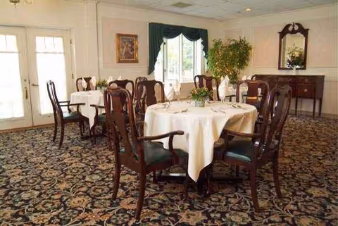 A dining room with round tables, white tablecloths, dark wood chairs, and a patterned carpet.