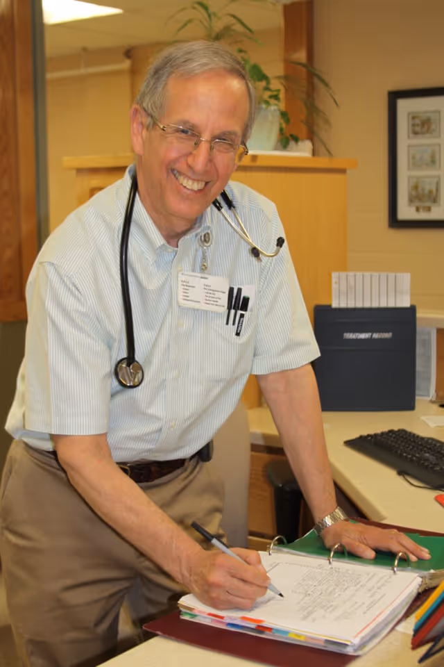 A smiling male healthcare professional wearing glasses, a light striped shirt, and a stethoscope around his neck is writing in a binder on a desk in an office setting. There are office supplies, a keyboard, and a treatment record folder visible on the desk behind him.