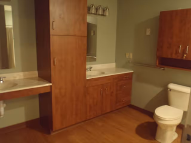 Interior view of a bathroom featuring a toilet, two sinks with countertops, wooden cabinets, a mirror, and a towel rack on the wall. The floor is wooden and the walls are painted green.