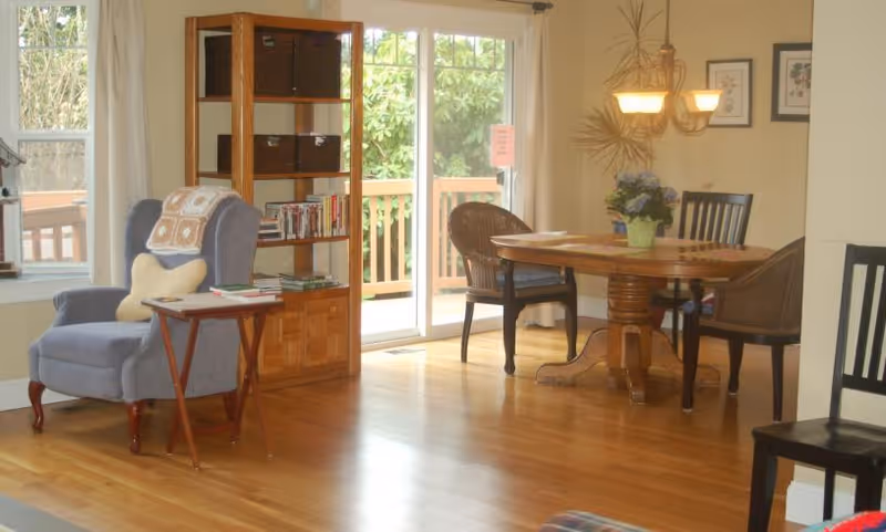 A cozy living and dining area with a blue armchair and small wooden side table on the left, a wooden bookshelf with baskets and books near a sliding glass door leading to an outdoor deck. On the right, a round wooden dining table with four chairs, a hanging light fixture above, and framed artwork on the wall. The room has hardwood floors and light-colored walls.