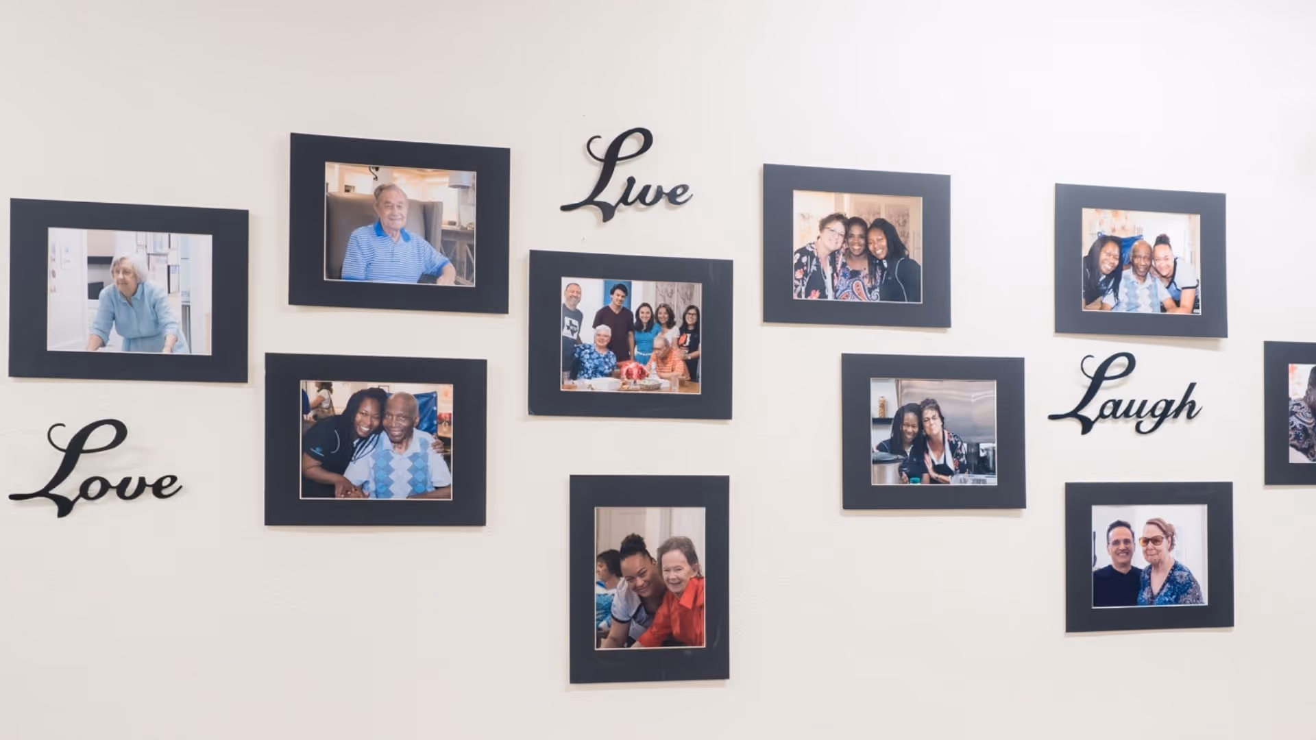 A wall decorated with multiple framed photos showing elderly residents and caregivers smiling and interacting warmly. The words 'Love', 'Live', and 'Laugh' are artistically displayed among the photos.