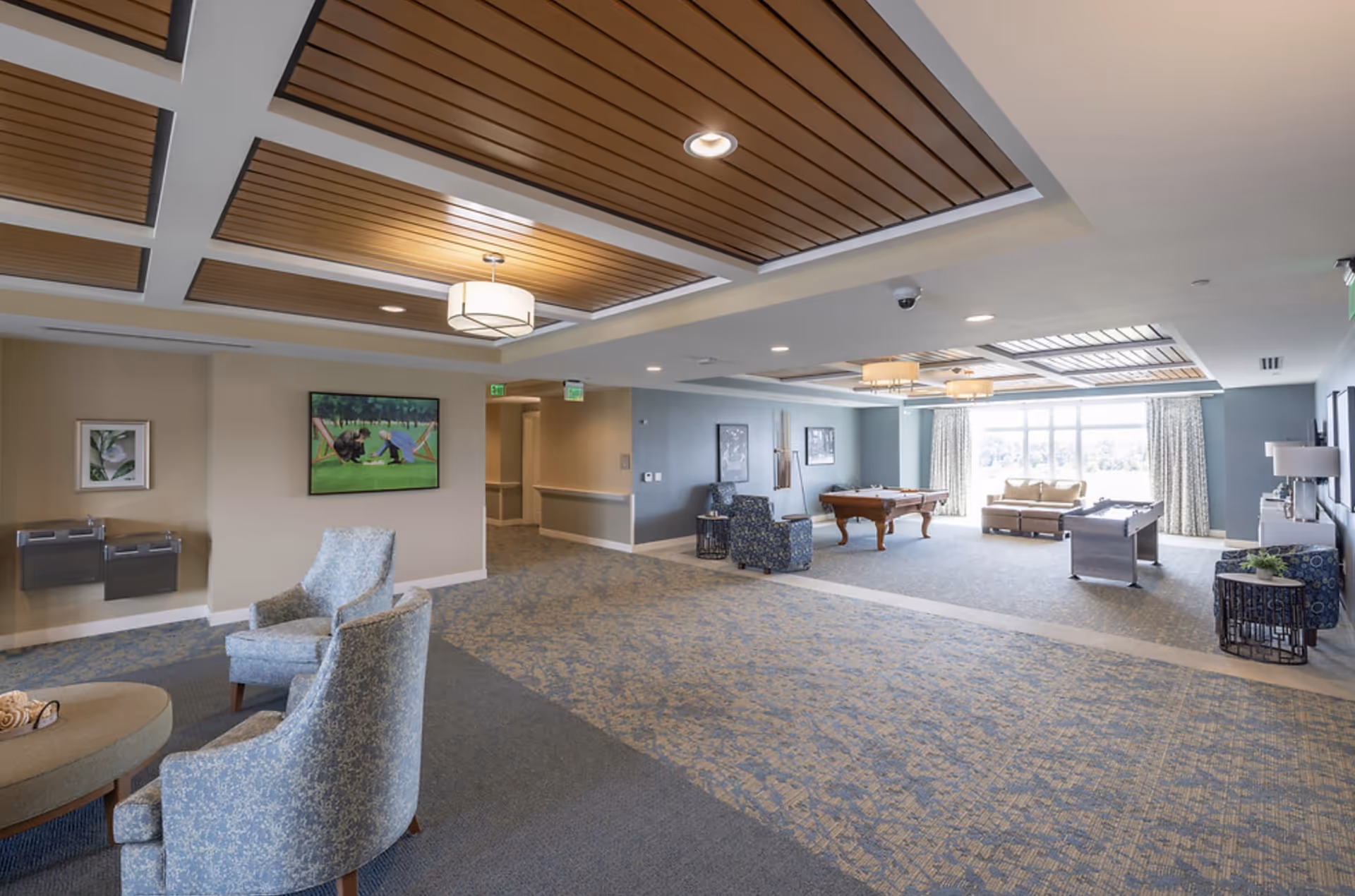 Spacious, well-lit senior living common room with comfortable seating, a pool table, game tables, and large windows.