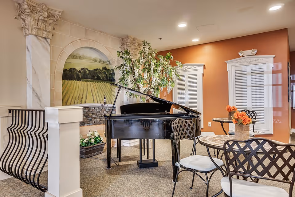 Interior common area with a black grand piano, metal chairs with white cushions, and round tables with flower arrangements. The walls are decorated with a mural of a vineyard and faux window shutters. There is a decorative plant near the piano and warm lighting overhead.