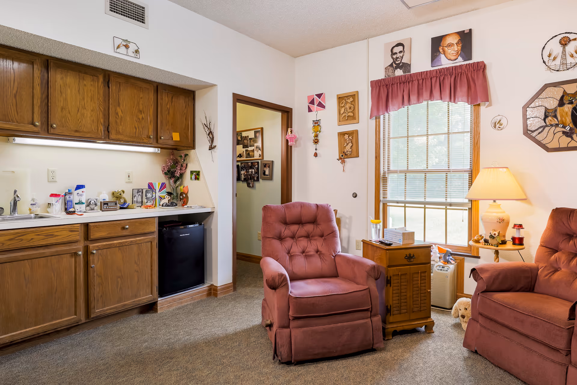 A cozy senior living room area with two pink upholstered armchairs, a wooden side table with a lamp, and a window with blinds and a pink valance. To the left, there is a kitchenette with wooden cabinets, a small black refrigerator, and various items on the countertop. The walls are decorated with framed pictures, artwork, and small hanging decorations.