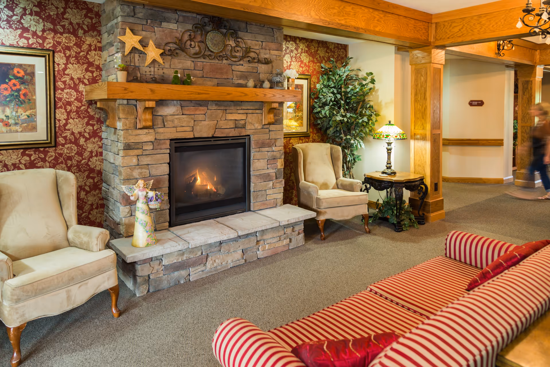 Cozy senior living community lounge area with a stone fireplace, two beige armchairs, a striped red and white sofa, a side table with a decorative lamp, floral wallpaper, and wooden beams. A person is walking in the background near a hallway.