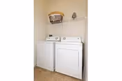 A small laundry room with a white washing machine and a white dryer side by side. Above them is a white wire shelf holding a wicker laundry basket. The floor is tiled in a light brown color.