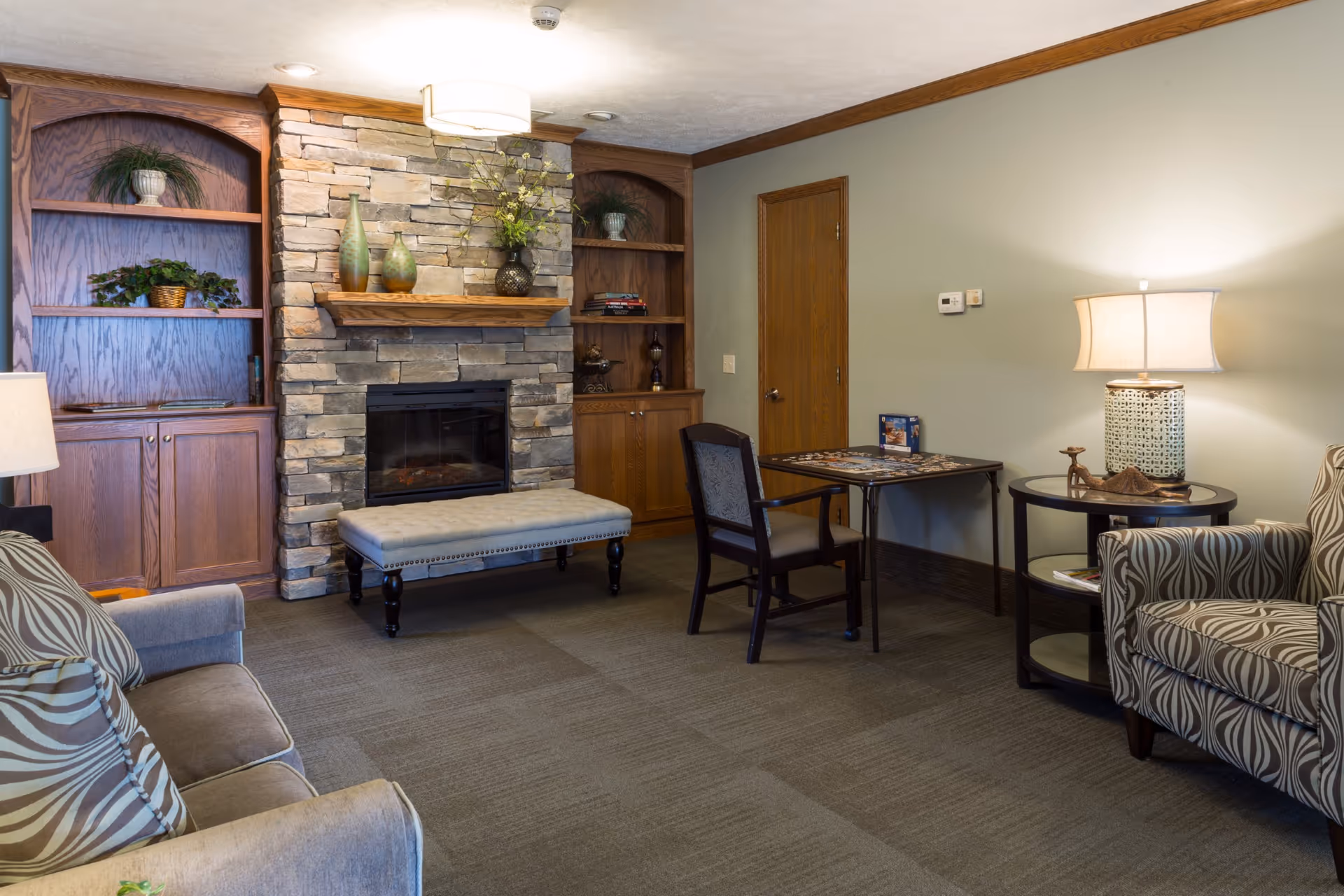 A cozy living room area featuring a stone fireplace with a wooden mantel decorated with vases and plants. On either side of the fireplace are built-in wooden shelves with decorative items. In front of the fireplace is a cushioned bench. To the right, there is a small table with a chair and a puzzle on it, next to a round side table with a lamp and decorative items. A patterned armchair and a sofa with striped pillows are also visible. The room has carpeted flooring and soft lighting.