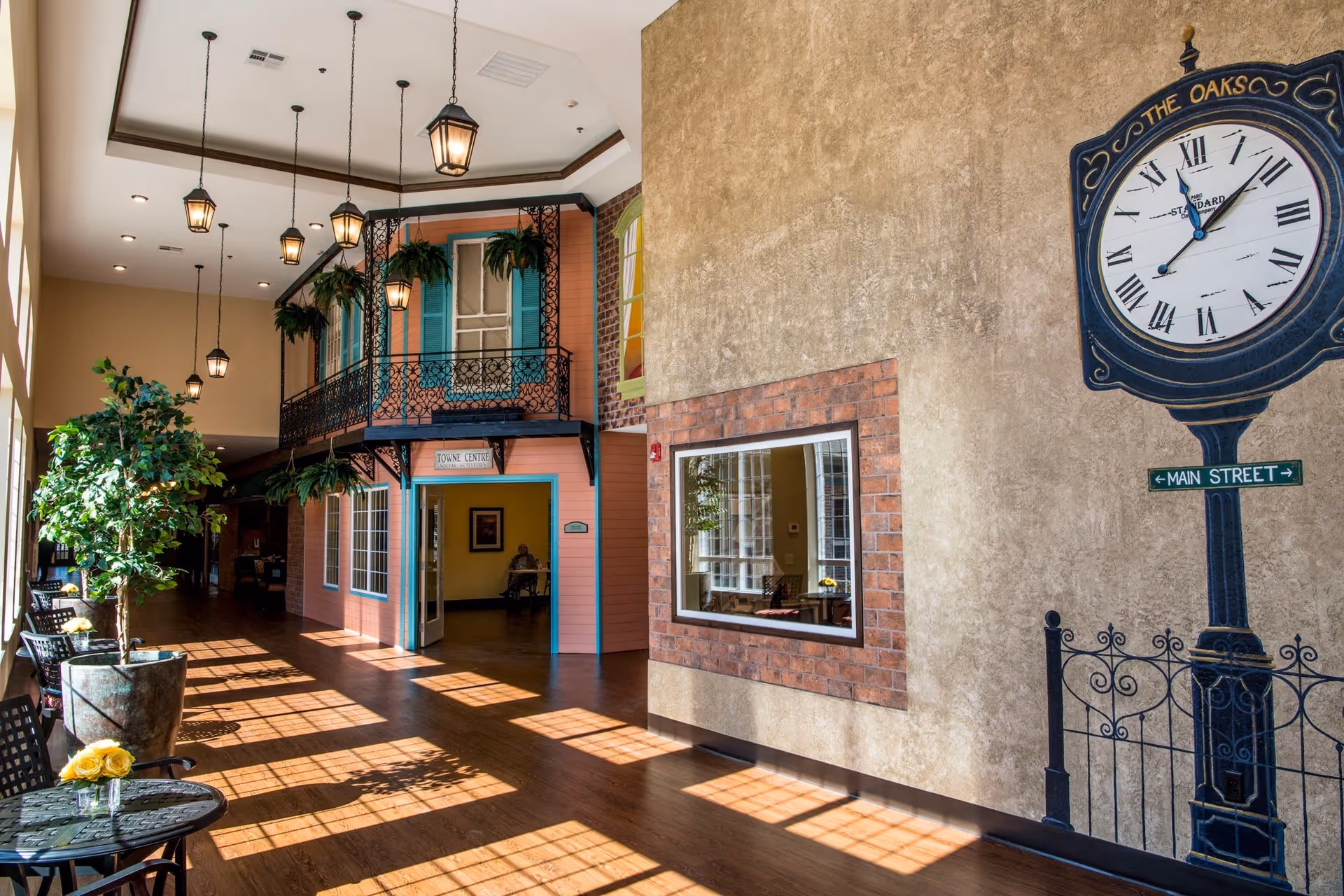 Interior hallway of The Oaks at Liberty Grove assisted living facility with wooden flooring, hanging lantern-style lights, potted plants, small tables with chairs, and a decorative wall clock showing 'The Oaks' and 'Main Street'. The hallway features a storefront-style facade labeled 'Towne Centre' with blue window shutters and hanging plants.