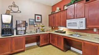 Interior view of a kitchen area with wooden cabinets, a microwave, a coffee machine, a water dispenser, and a countertop with a sink. The walls are painted in beige and red tones, and there is a decorative coffee cup wall art above the coffee machine.