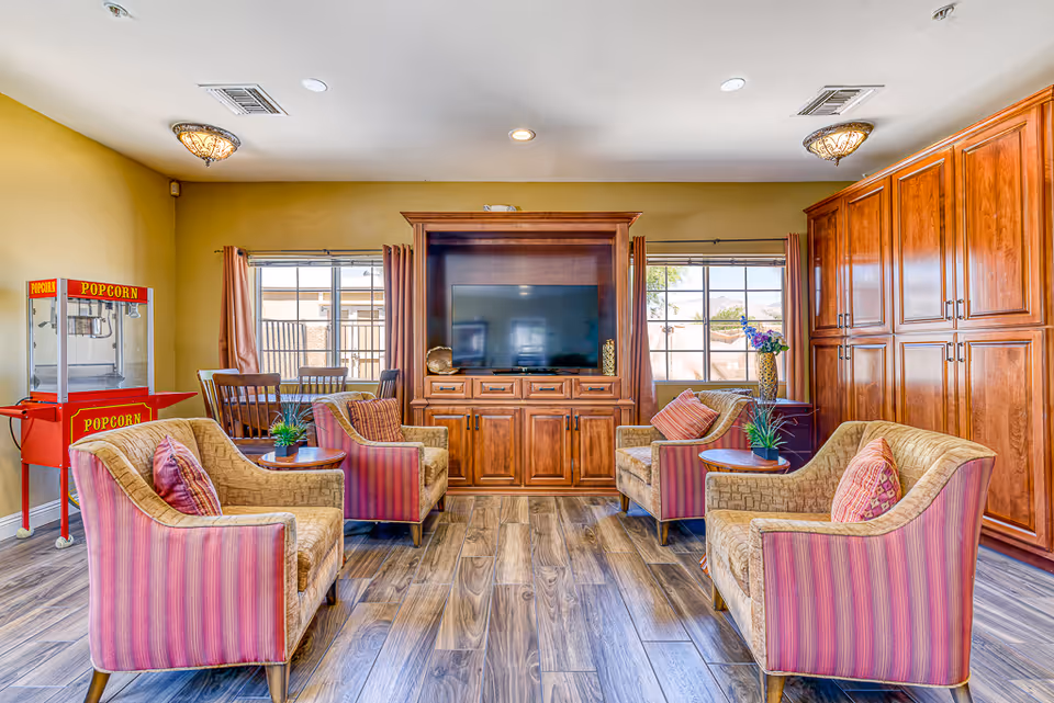 A bright common sitting area with upholstered armchairs arranged around a central wooden TV cabinet, large windows, built-in cabinetry, and a red popcorn machine.