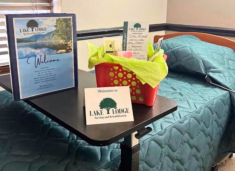 A small table between two beds in a nursing facility room. On the table is a red basket with green tissue paper and various items inside, along with a welcome card and a framed welcome sign for Lake Lodge Nursing and Rehabilitation. The beds have teal quilted bedspreads.