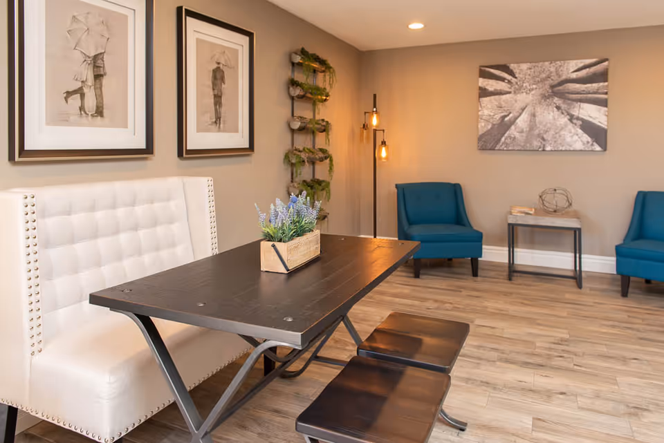 A cozy interior room featuring a dark wooden table with a small rectangular planter of purple flowers on top. The table is accompanied by a white cushioned bench with nailhead trim on one side and two dark wooden stools on the other. In the background, there are two blue upholstered chairs, a small side table with decorative items, a tall floor lamp with exposed bulbs, and wall art including two framed pictures of people with umbrellas and a large black and white photograph of tree trunks.
