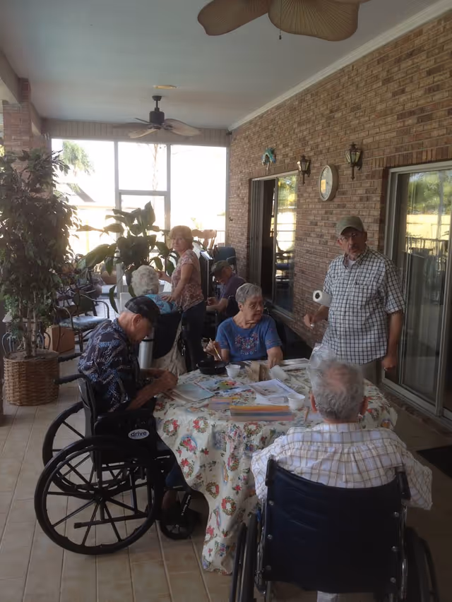 Elderly residents gathered around a floral-covered table on a shaded, screened porch with plants and ceiling fans.