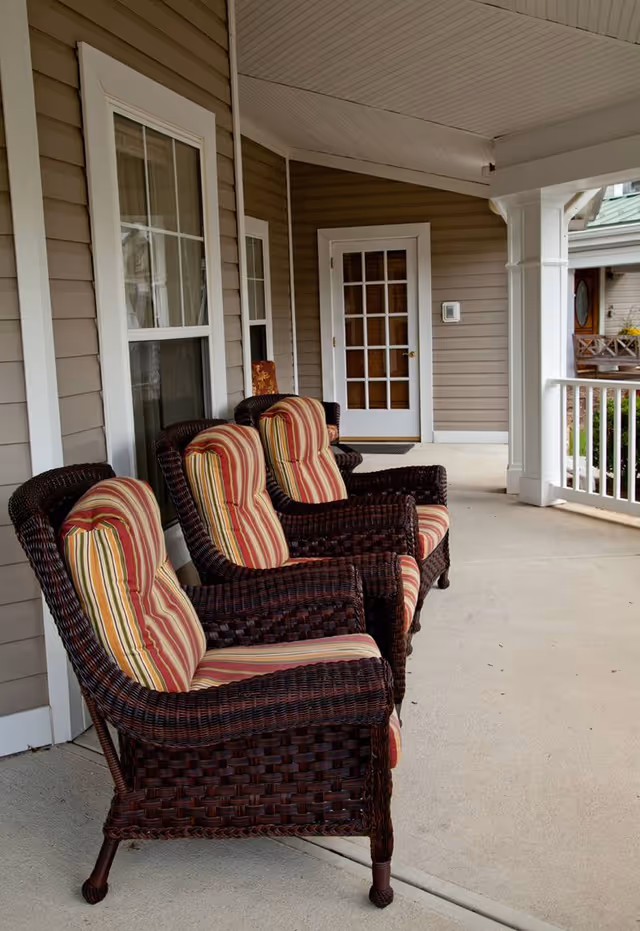A covered porch area with four wicker chairs featuring striped cushions in red, yellow, and beige. The porch has beige siding, white trim around windows and doors, and a white railing. A glass-paneled door is visible at the end of the porch.