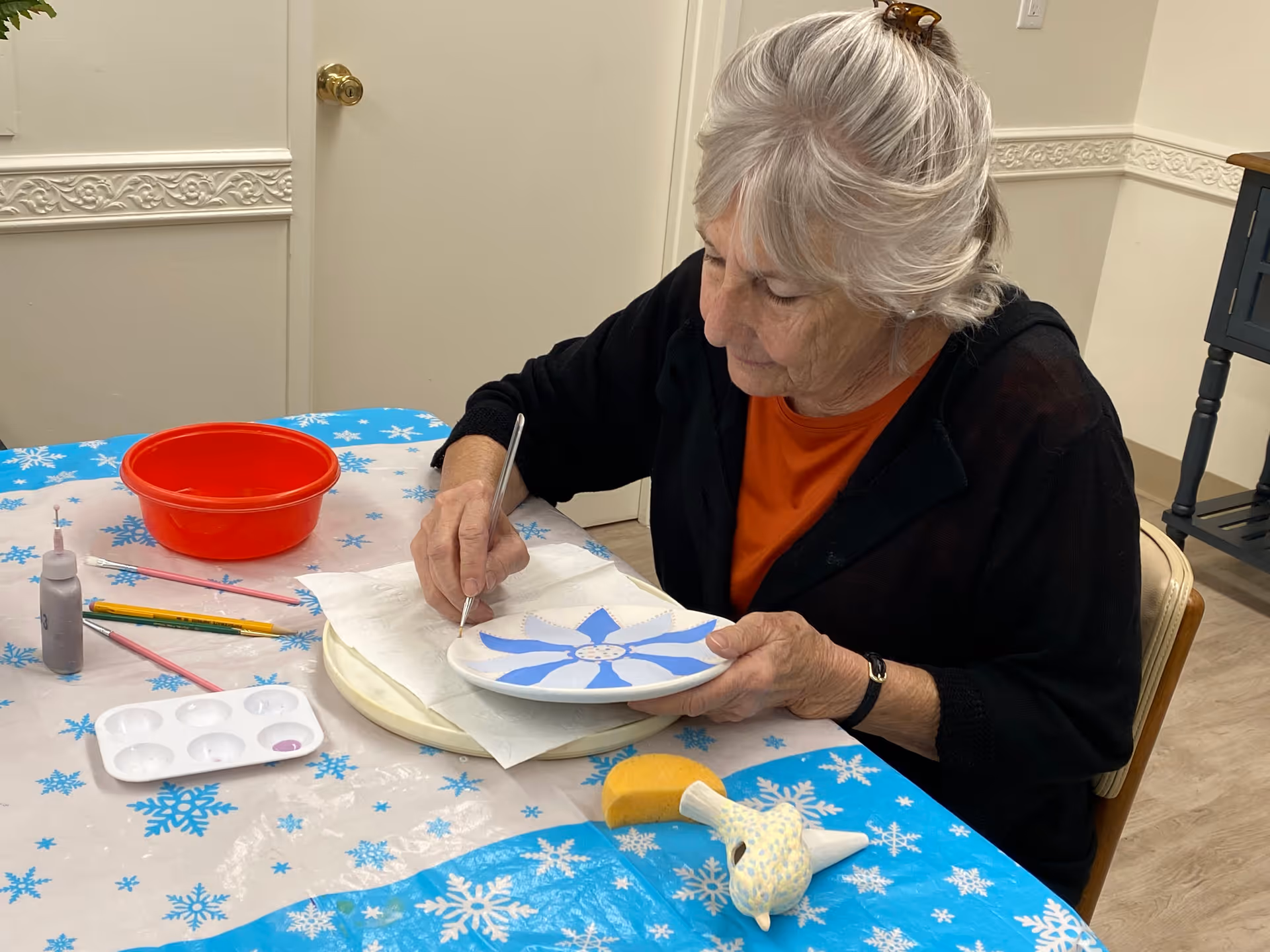 An elderly woman with gray hair is seated at a table covered with a snowflake-patterned tablecloth. She is painting a white plate with blue petal-like designs using a fine brush. On the table are various painting supplies including a red bowl, paintbrushes, a paint palette, and a small yellow sponge with a bird-shaped handle. The room has light-colored walls with decorative molding and a wooden floor.