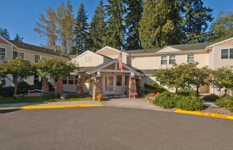 Front exterior view of a senior living facility building with beige siding, stone pillars at the entrance, an American flag above the door, surrounded by trees and landscaping under a clear blue sky.