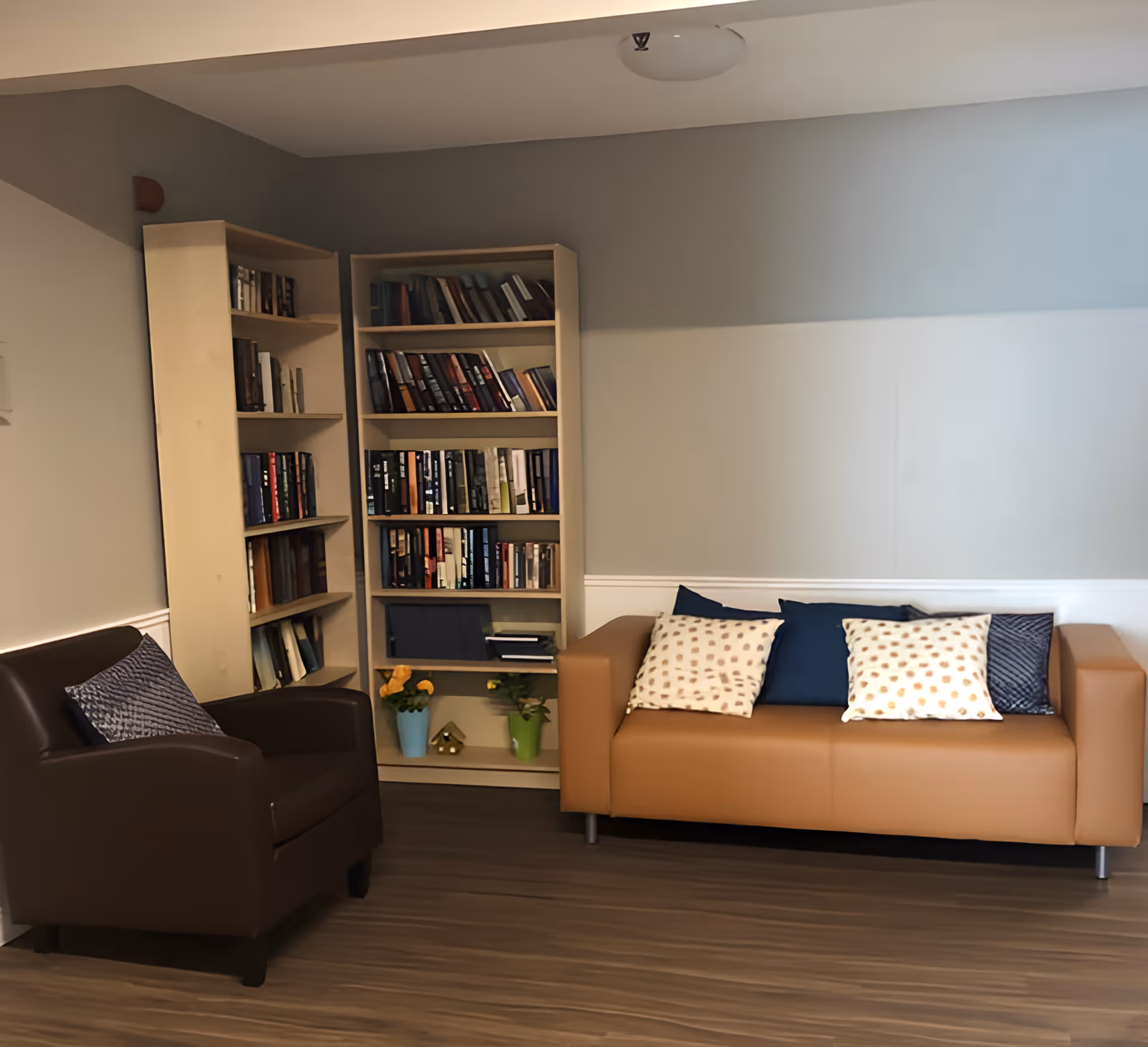 A lounge area with a tan sofa and brown armchair beside a bookshelf filled with books.