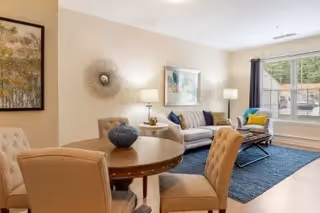 A bright living room with a round wooden dining table and four upholstered chairs in the foreground. The room features a beige sofa with colorful cushions, a coffee table on a blue rug, two table lamps, a large window with dark curtains, and wall art including a framed painting and a decorative wall sculpture.