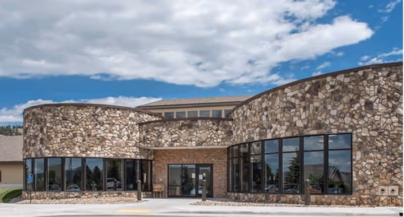 Front exterior view of Fountain Springs Healthcare Center featuring a building with a stone facade, large windows, and a clear blue sky with some clouds above.