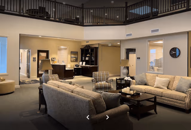A spacious and well-lit living room area in a senior living facility with beige sofas, a plaid armchair, a wooden coffee table with candles, two table lamps, and a dark wood cabinet in the background. The room has a high ceiling with a balcony railing above and neutral-colored walls.