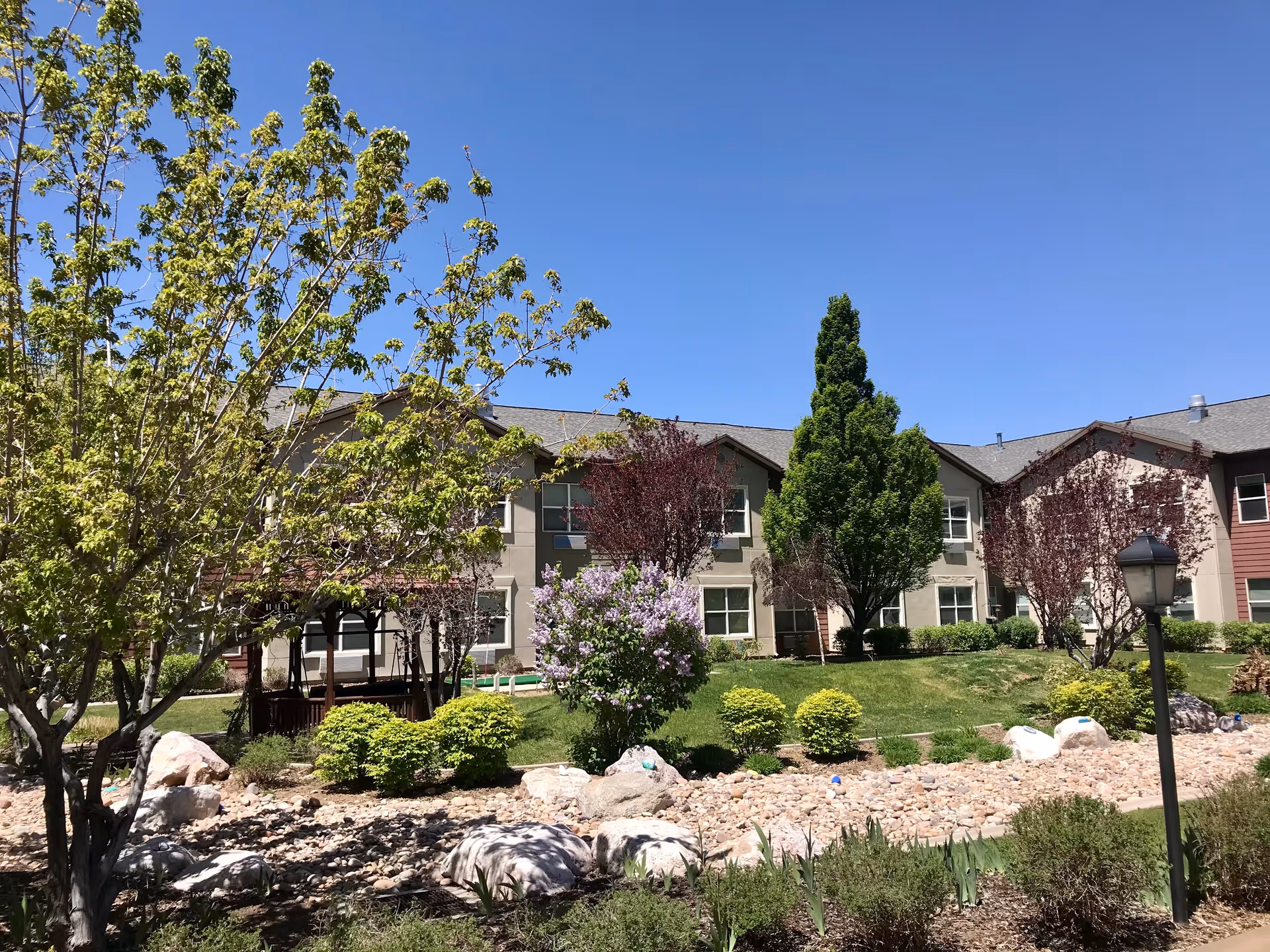 Two-story senior living building front with landscaped lawn, trees, shrubs and decorative rocks beneath a clear blue sky.