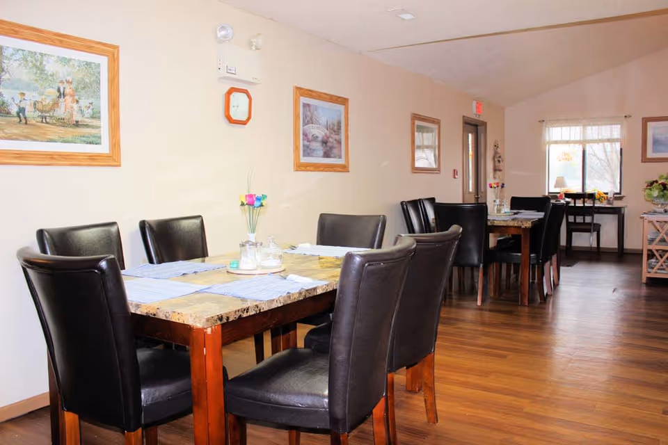 Interior view of a dining room with two wooden tables topped with marble-like surfaces, each surrounded by black leather chairs. The room has wooden flooring, light-colored walls adorned with framed paintings, and a window letting in natural light. There are placemats and a small flower vase on the tables.