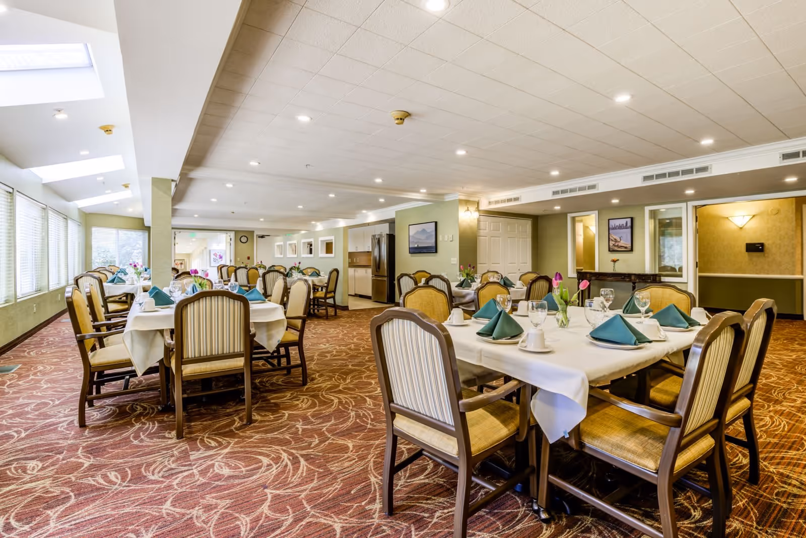 A spacious dining room with multiple tables set for a meal, each table covered with white tablecloths, green folded napkins, glassware, and small flower arrangements. The room has large windows on one side allowing natural light, patterned carpet flooring, and soft ceiling lighting.