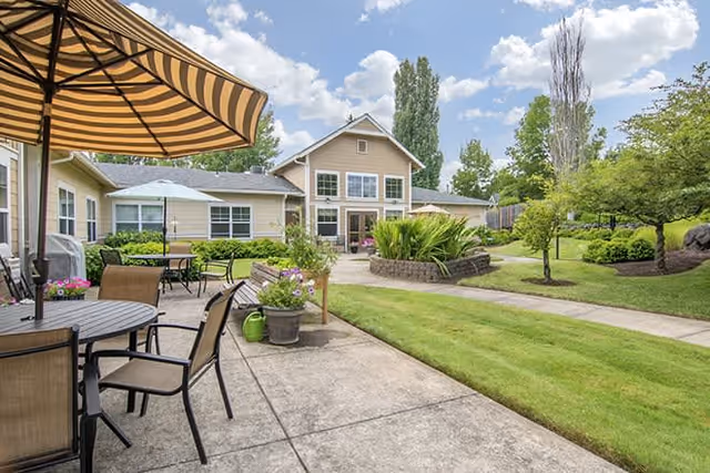 Outdoor patio area with tables, chairs, and a large striped umbrella next to a well-maintained lawn and garden. In the background, there is a beige building with multiple windows and a clear blue sky with some clouds.