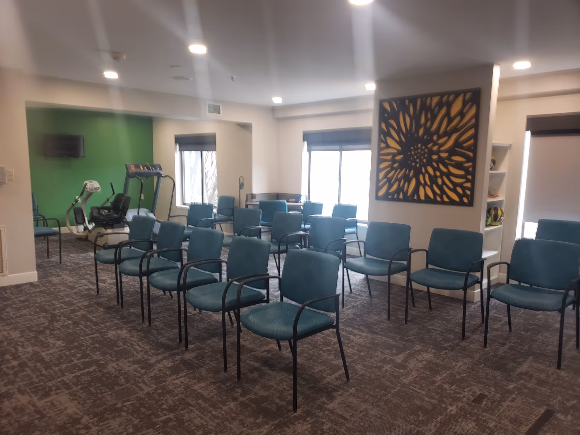 A room with multiple teal chairs arranged in rows facing forward, with exercise equipment including a treadmill and stationary bike visible in the background. The room has carpeted flooring, white walls, recessed ceiling lights, and a large decorative wall art piece with a floral pattern. There are windows with blinds partially drawn, and a small shelving unit with sports balls on the right side.
