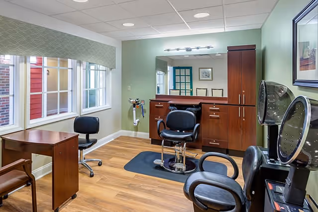 A well-lit salon room with wooden flooring and light green walls. The room contains salon chairs, a wooden cabinet with a large mirror, a small desk, and two hair drying stations. There are three windows with a patterned valance allowing natural light into the room.