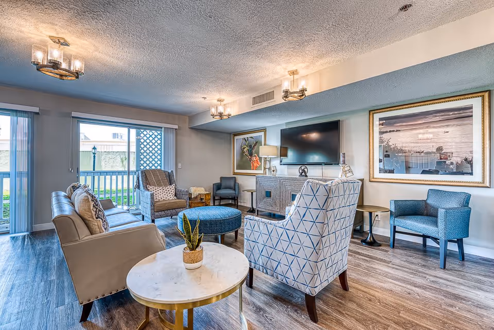 Bright furnished lounge with sofas, patterned armchairs, coffee tables, a wall-mounted TV, and framed artwork.