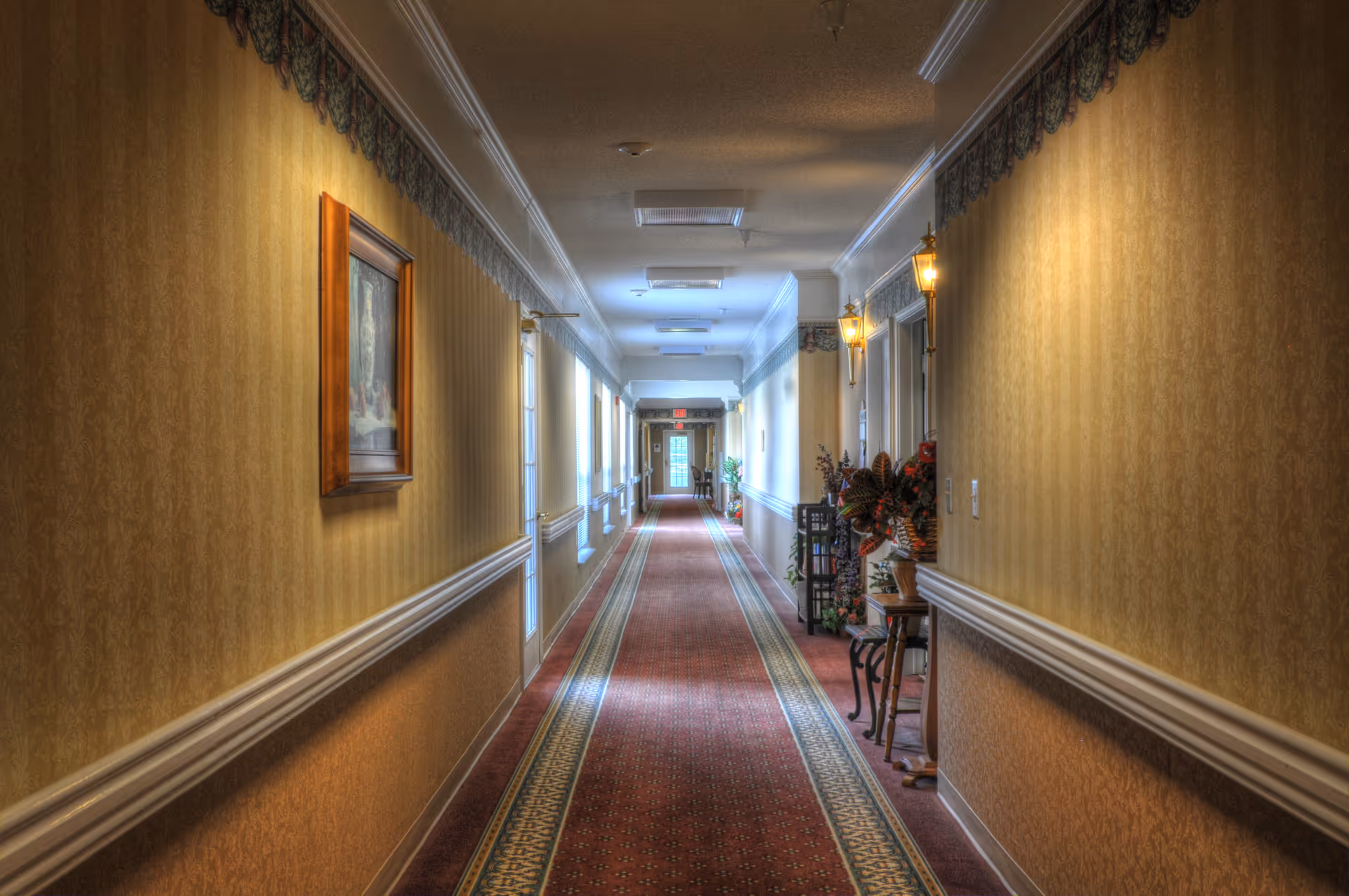 Long carpeted interior hallway with patterned wallpaper, wall sconces, framed art and decorative tables.