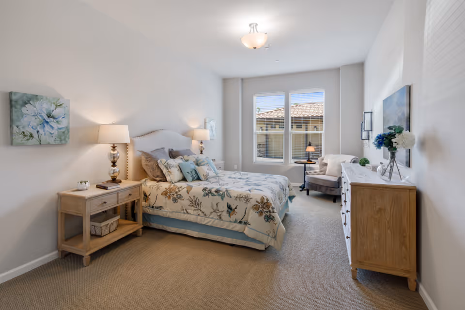A bright and cozy bedroom with a large bed adorned with floral bedding and multiple pillows. There are two bedside tables with lamps, a wooden dresser with decorative items including a vase with flowers, and a comfortable armchair near two large windows letting in natural light. The walls are painted white and decorated with floral artwork.