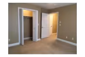 Empty room with beige walls and carpeted floor, featuring an open closet with white bi-fold doors and a white door leading to another room or hallway.