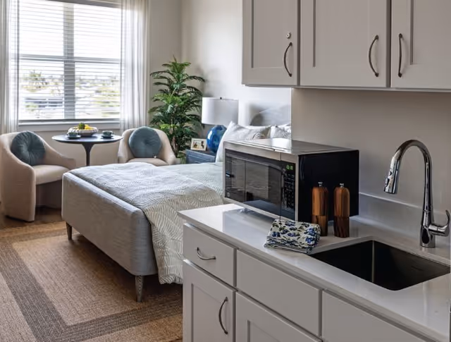 A cozy studio apartment featuring a bed with a light-colored bedspread, two cushioned chairs with round pillows, a small round table with fruit, a potted plant, a bedside table with a lamp and picture frame, and a kitchenette area with white cabinets, a microwave, a sink, and a faucet.
