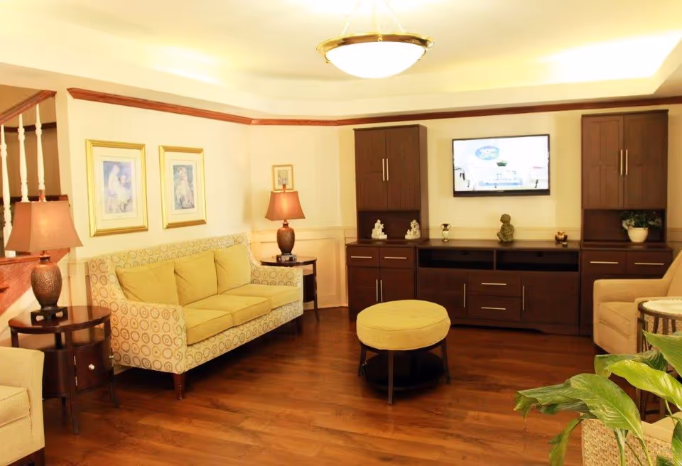 Cozy common living area with a patterned sofa, armchairs, central ottoman, wall-mounted TV and wooden cabinets.