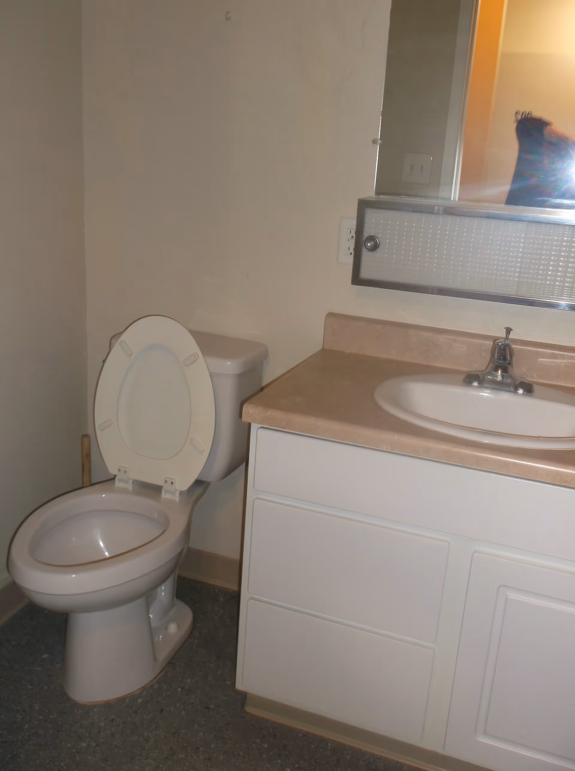 A small bathroom with a white toilet with the lid open, a beige countertop with a white sink, and a mirror above the sink. The walls are plain and light-colored, and the floor has a speckled pattern.