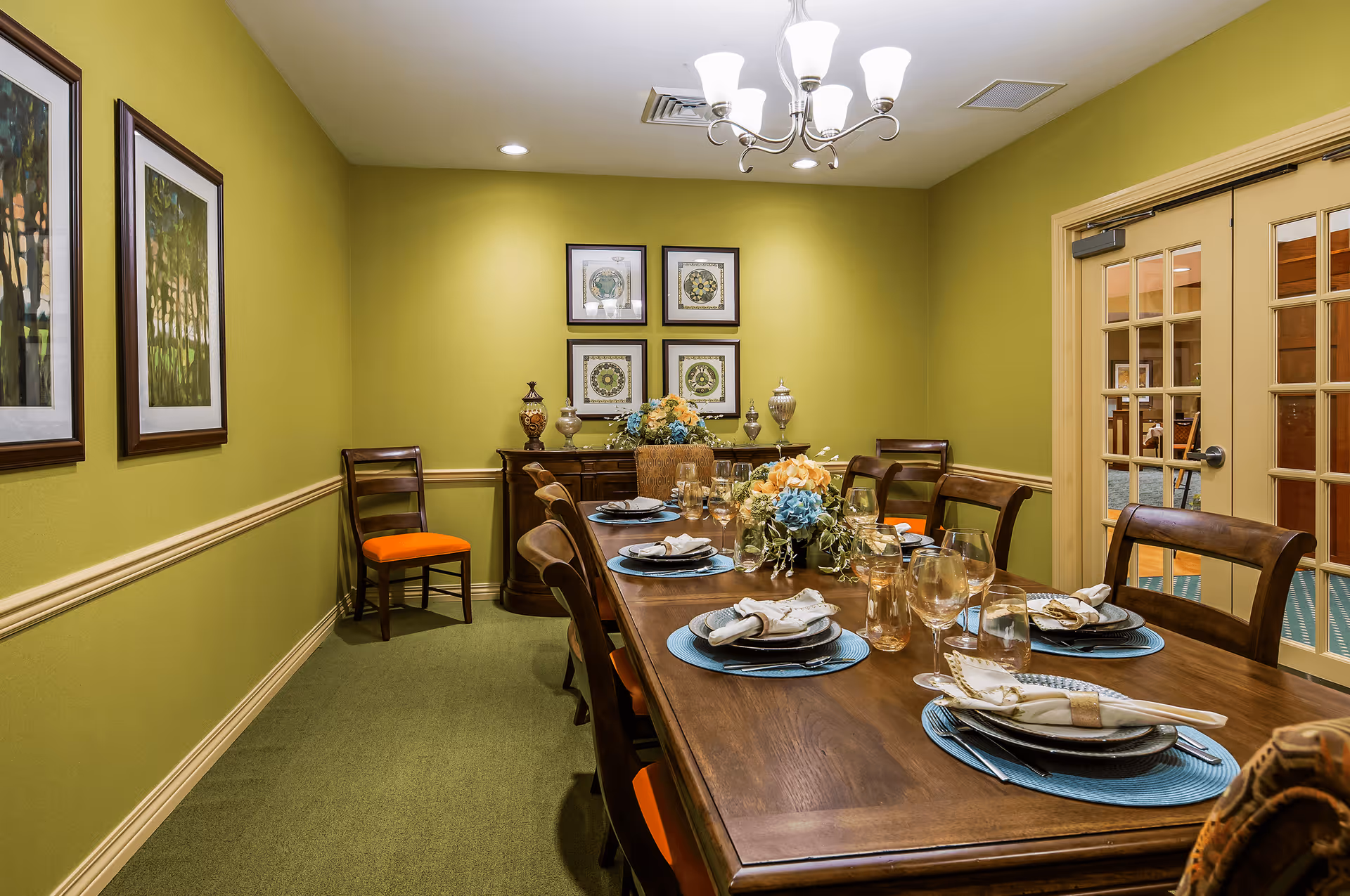 A formal dining room with a long wooden table set for a meal. The table has blue placemats, plates with napkins, glassware, and floral centerpieces. The walls are painted green with framed artwork and decorative vases on a sideboard. There are wooden chairs with orange cushions around the table, and a chandelier with multiple lights hangs from the ceiling. A set of glass-paneled double doors is visible on the right side of the room.